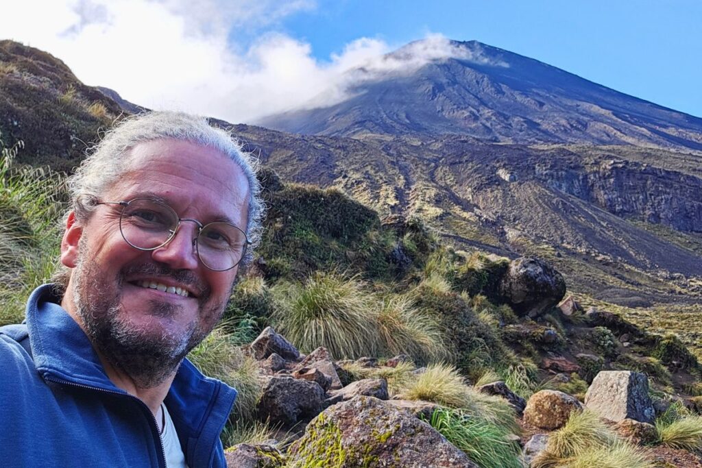 Reiseblogger Nicolo Martin lächelt vor dem kegelförmigen Vulkan Mount Ngauruhoe (Schicksalsberg) im Tongariro Nationalpark unter blauem Himmel.