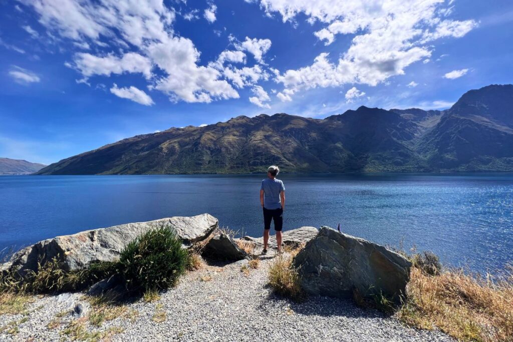 Nicolo Martin blickt von Felsen aus auf einen weiten, tiefblauen See und die Bergkette der Südinsel unter strahlender Sonne.
