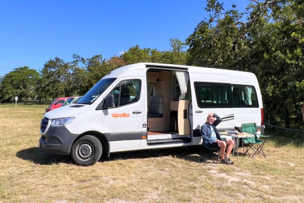 Reiseblogger Nicolo sitzt entspannt beim Frühstück vor seinem weißen Apollo-Campervan auf einem grünen Stellplatz in Neuseeland.