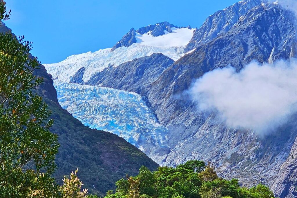 Der blau schimmernde Franz Josef Gletscher fließt zwischen steilen Bergwänden im Westland Nationalpark hinab, teilweise von Wolken verdeckt.