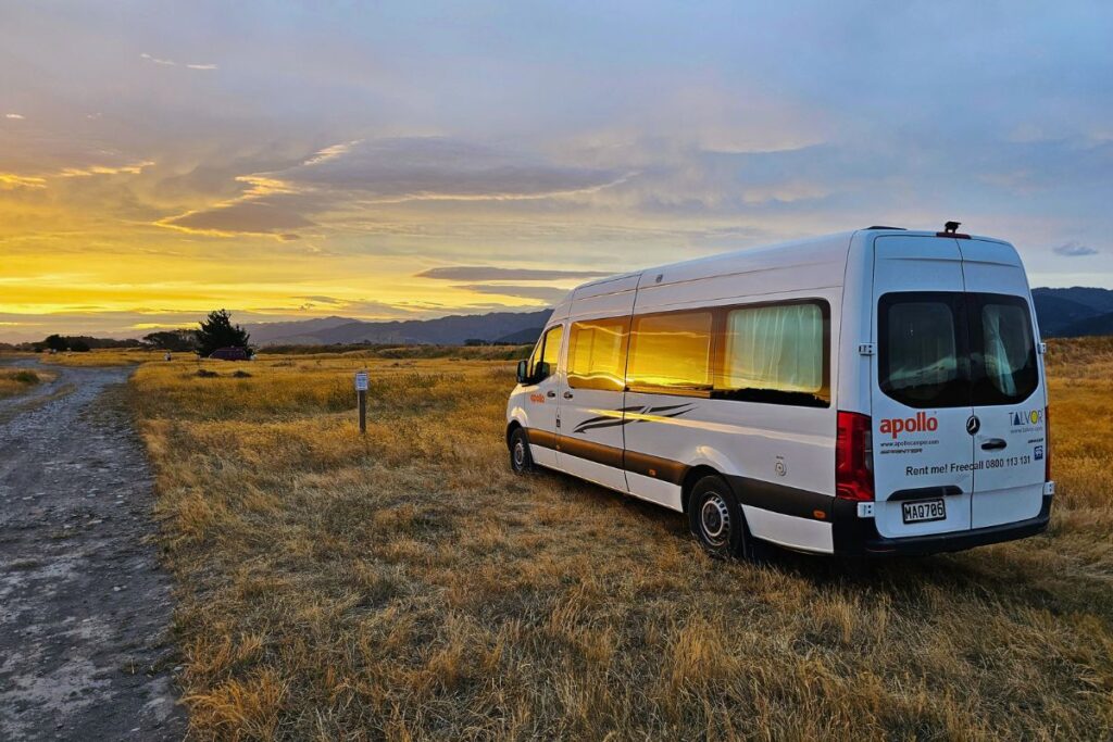 Ein weißer Apollo Campervan steht bei einem goldenen Sonnenuntergang auf einer weiten Graslandschaft in Neuseeland.