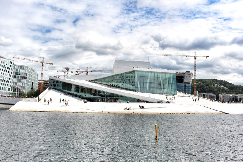 Panoramablick auf das weiße Opernhaus in Oslo (Operaen) vom Fjord aus gesehen. Menschen spazieren auf dem schrägen Marmordach, im Hintergrund sind Baukräne und der blaue Himmel mit Wolken zu sehen.