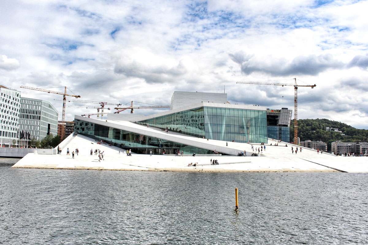 Panoramablick auf das weiße Opernhaus in Oslo (Operaen) vom Fjord aus gesehen. Menschen spazieren auf dem schrägen Marmordach, im Hintergrund sind Baukräne und der blaue Himmel mit Wolken zu sehen.