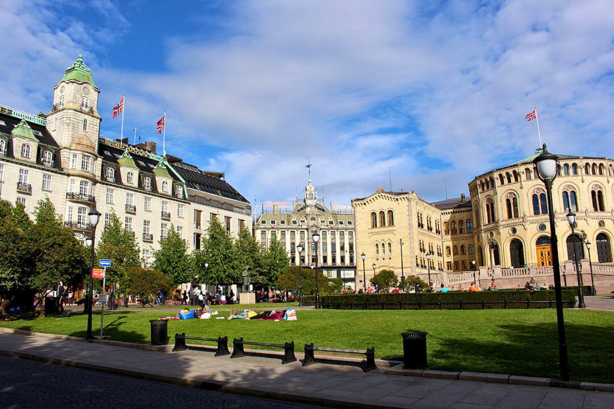 Panoramablick auf den Eidsvolls plass in Oslo: Links das Grand Hotel, rechts das Parlament (Stortinget), dazwischen grüner Park unter blauem Himmel
