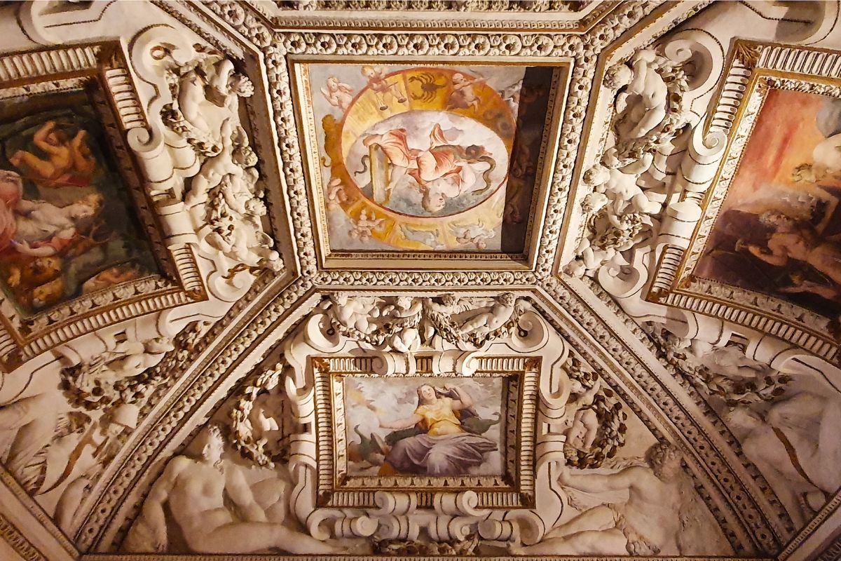 Nahaufnahme einer prunkvoll verzierten Decke im Palazzo Thiene mit weißen Stuckfiguren und Malereien, die astrologische Motive und Sternzeichen zeigen.