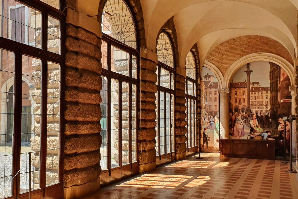 elle Galerie im Palazzo Thiene mit hohen Bogenfenstern auf der linken Seite und einem großen, farbenfrohen Wandgemälde am Ende des Ganges, das eine belebte Marktszene zeigt.