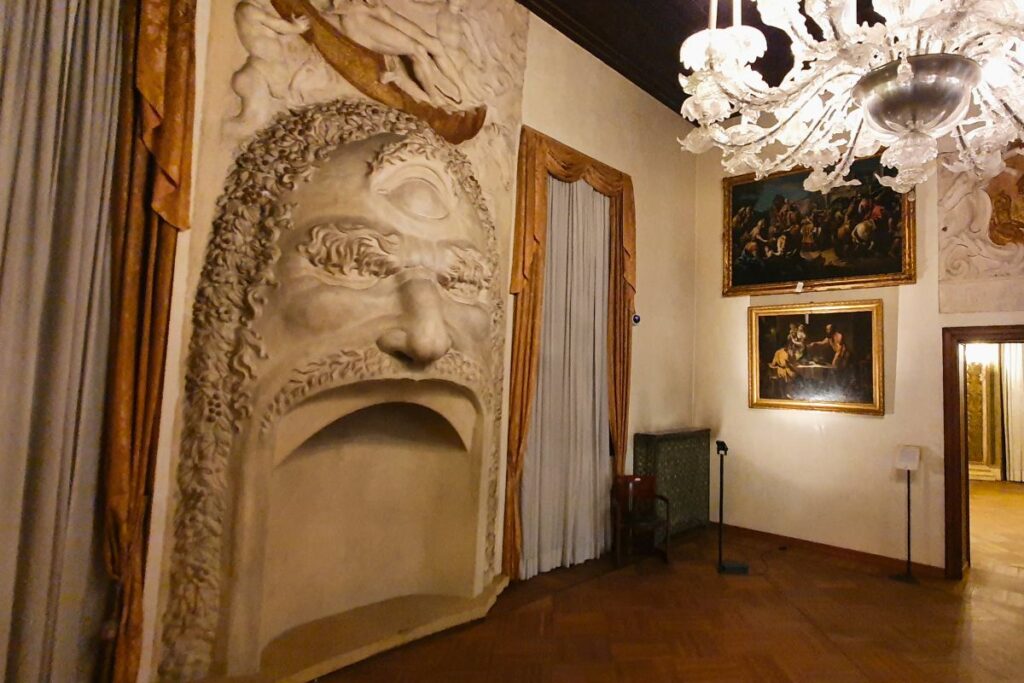 Ein außergewöhnlicher Kamin im Palazzo Thiene, gestaltet als riesiges, weißes Monstergesicht mit aufgerissenem Mund als Feuerstelle, daneben hängen klassische Ölgemälde an der Wand.
