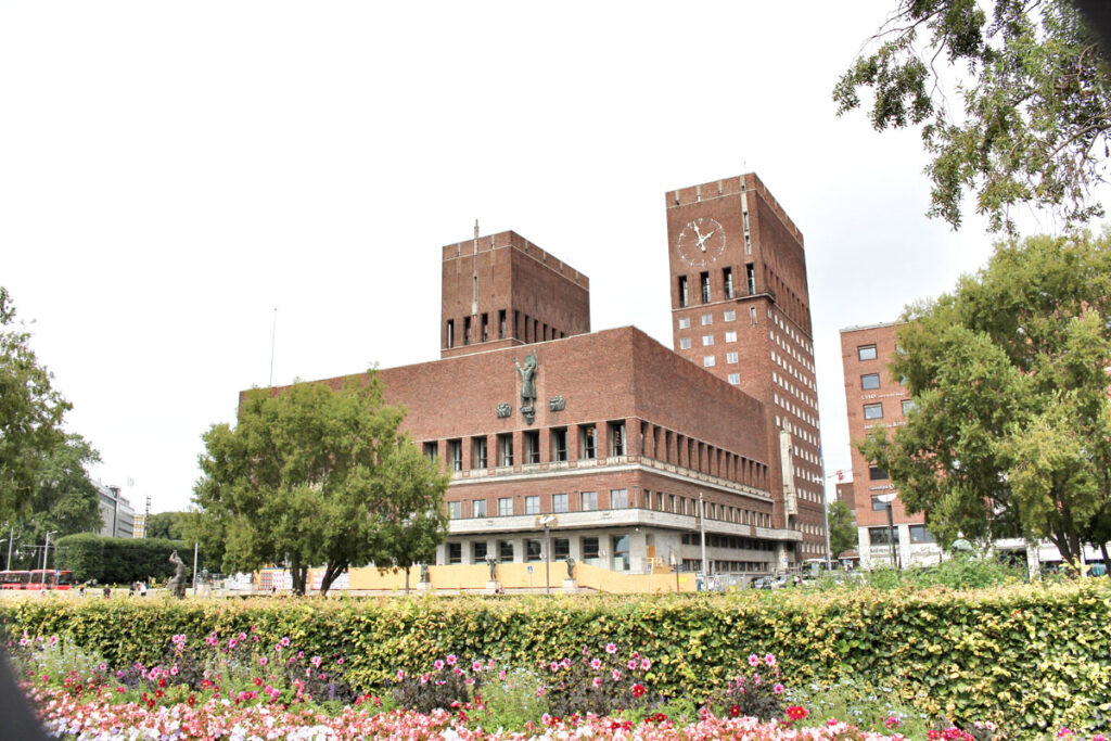 Das markante rote Backsteingebäude des Osloer Rathauses mit seinen zwei Türmen, davor bunte Blumenbeete und Touristen