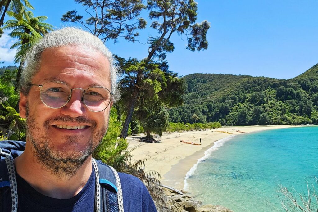 Reiseblogger Nicolo Martin lächelt glücklich in die Kamera, im Hintergrund erstreckt sich eine einsame Bucht mit türkisfarbenem Wasser im Abel Tasman Nationalpark.