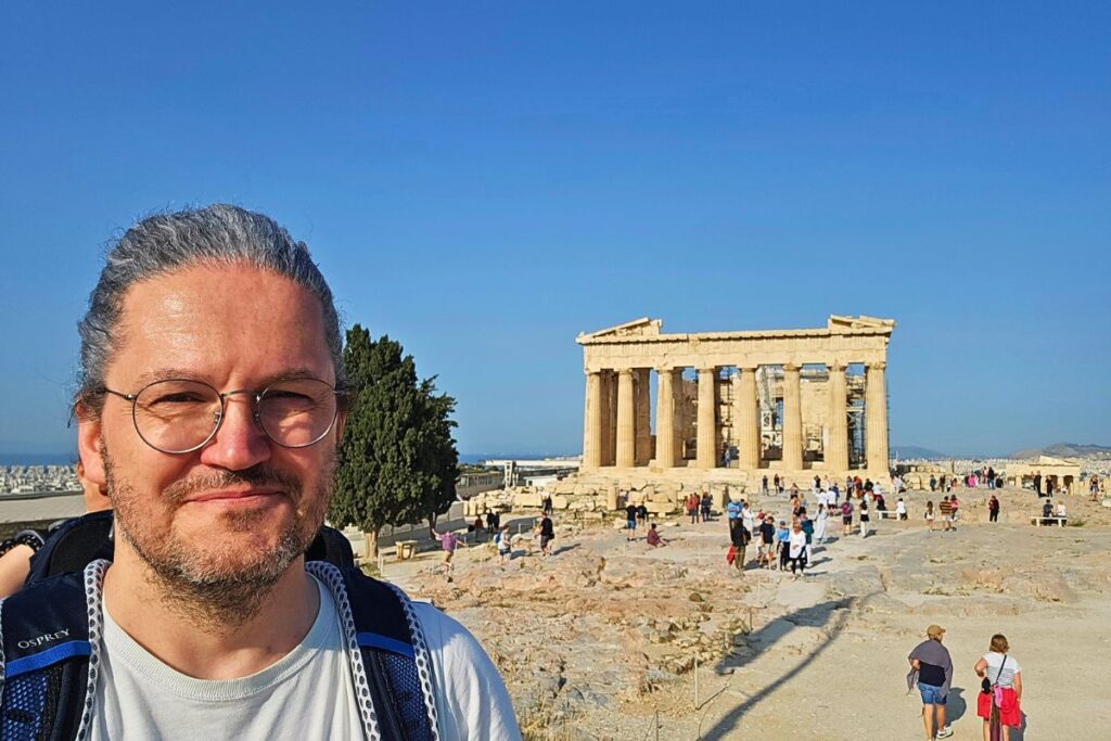 Reiseblogger Nicolo Martin lächelt glücklich vor der Kulisse des antiken Parthenon-Tempels auf der Akropolis in Athen.