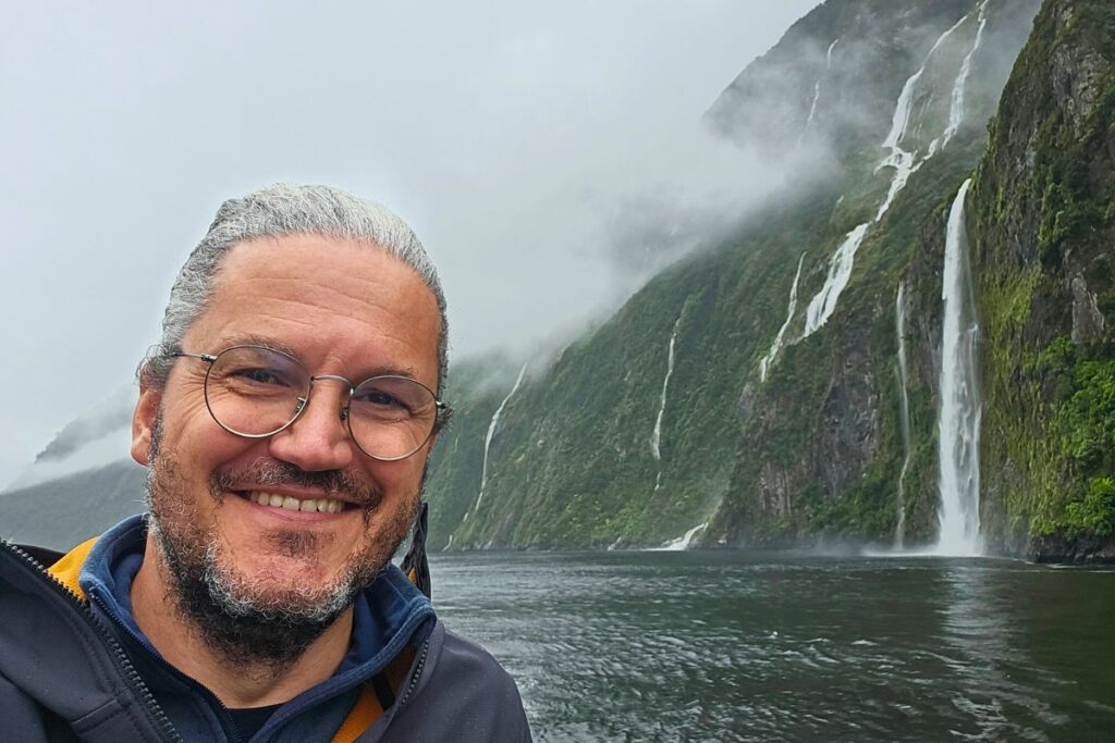 Reiseblogger Nicolo Martin lächelt bei regnerischem Wetter auf einem Boot im Milford Sound, im Hintergrund sind mehrere Wasserfälle an den grünen Bergen zu sehen.