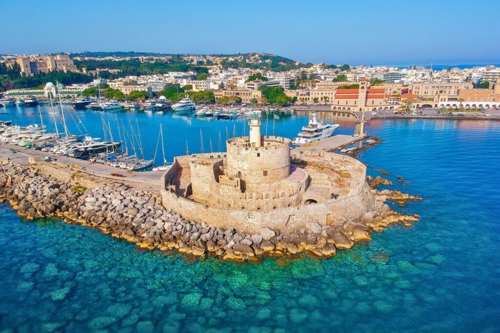 Luftaufnahme vom Mandraki-Hafen in Rhodos-Stadt mit der kreisförmigen Festung St. Nikolaus auf einer Mole, umgeben von türkisblauem Wasser und Segelyachten.