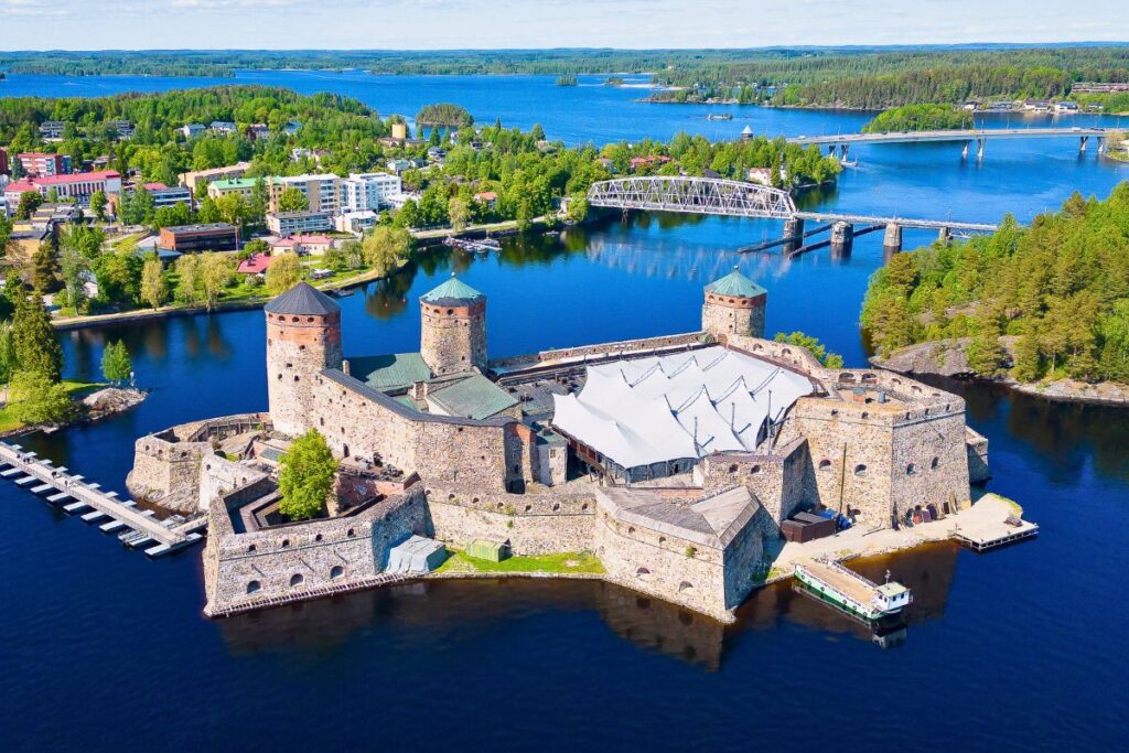 Luftaufnahme der mittelalterlichen Burg Olavinlinna in Savonlinna, umgeben von blauem Wasser und grünen Inseln.