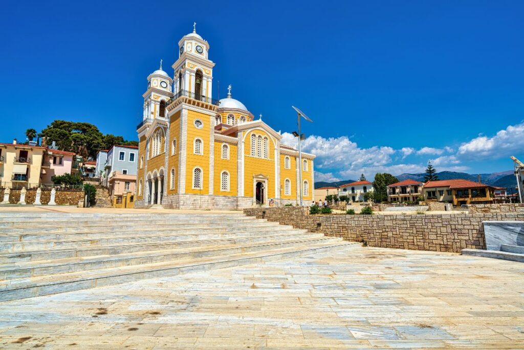 Die große, gelb-weiße Kathedrale Ypapanti in Kalamata auf dem Peloponnes vor einem weiten Marmorplatz unter strahlend blauem Himmel.