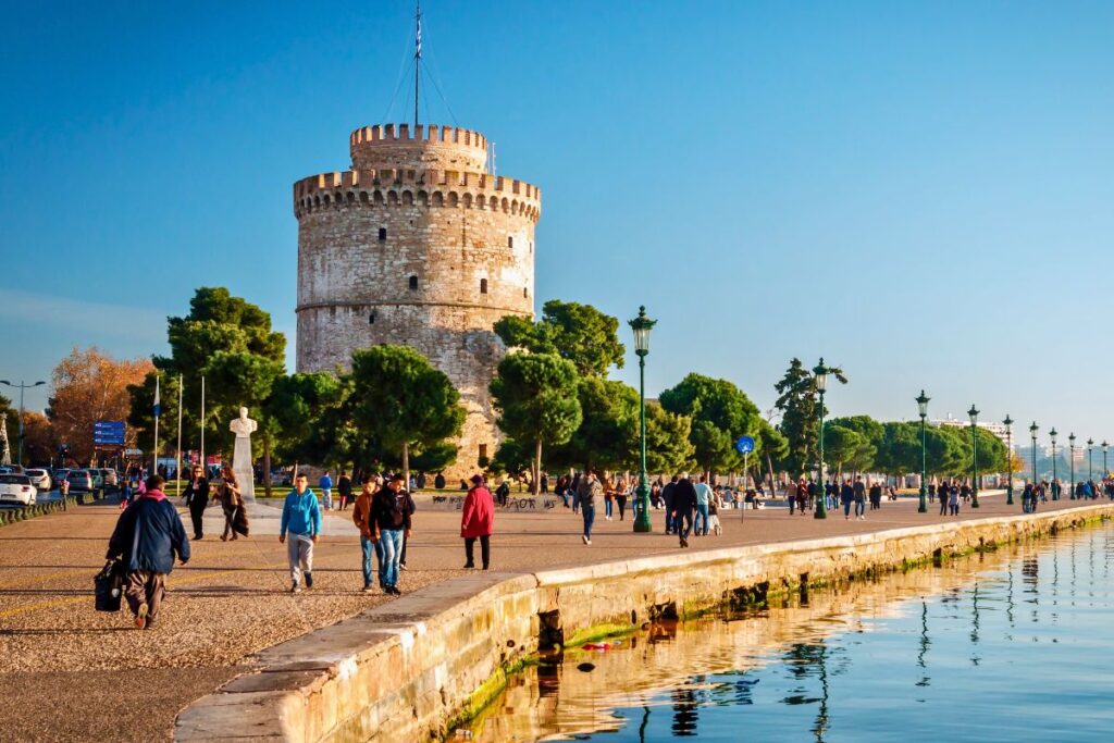 Der historische Weiße Turm von Thessaloniki an der sonnigen Uferpromenade, im Hintergrund Menschen beim Spaziergang unter blauem Himmel.