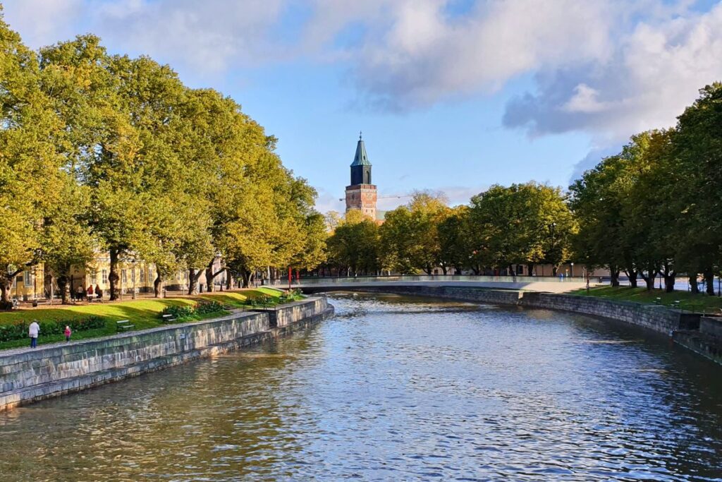 Der Fluss Aurajoki in Turku, gesäumt von grünen Bäumen und Spazierwegen, mit dem Turm des Doms im Hintergrund.