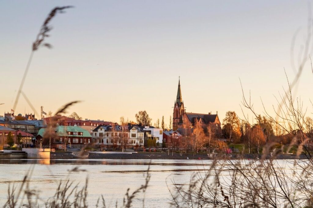 Blick über den breiten Fluss Ume älv auf die Stadtkirche von Umeå, die im warmen Licht des Sonnenuntergangs leuchtet.