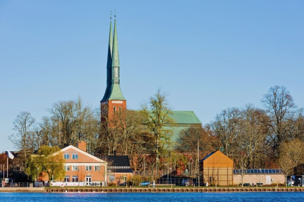 Der Dom zu Växjö mit seiner roten Fassade und den zwei spitzen Türmen, gesehen vom Ufer des Växjösjön-Sees an einem sonnigen Tag.