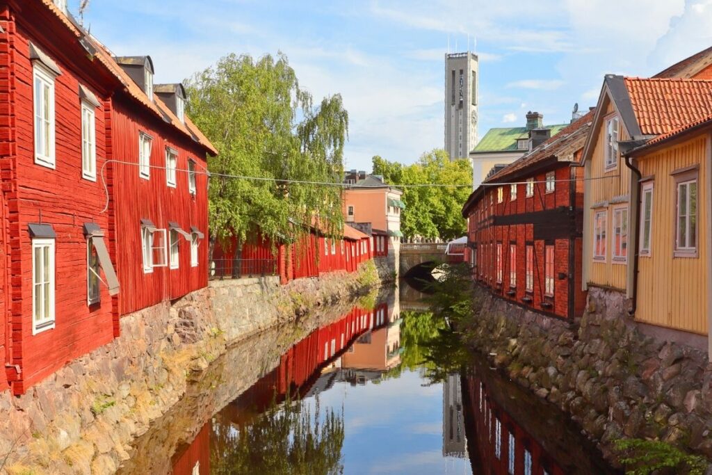 Rote historische Holzhäuser im Viertel Kyrkbacken in Västerås säumen einen schmalen Wasserlauf, im Hintergrund ragt der Turm des Doms auf.