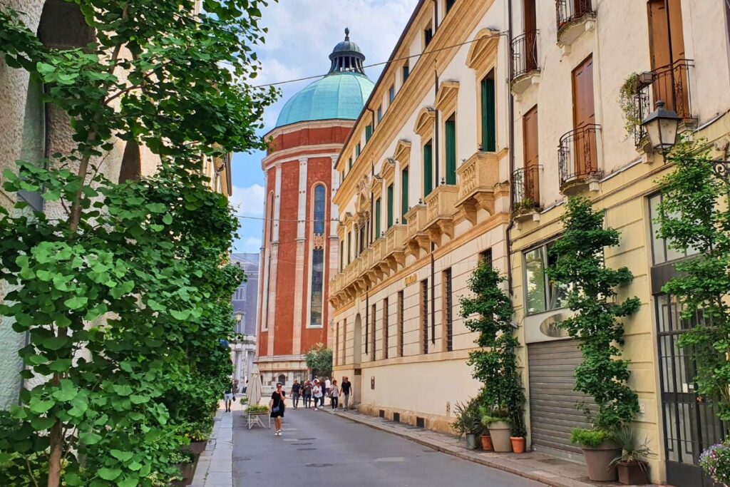 Blick durch eine schmale Gasse mit historischen Palästen auf die riesige, grüne Kuppel und die rote Apsis des Doms von Vicenza.