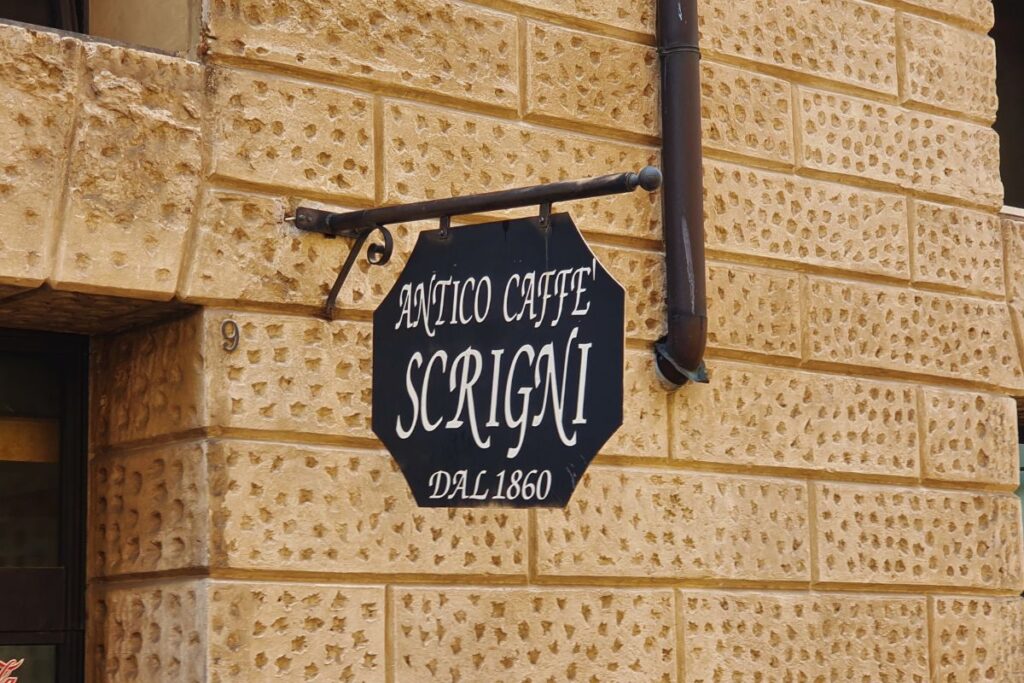Ein schwarzes, achteckiges Hängeschild an einer gelben Steinwand mit der Aufschrift "Antico Caffè Scrigni DAL 1860".