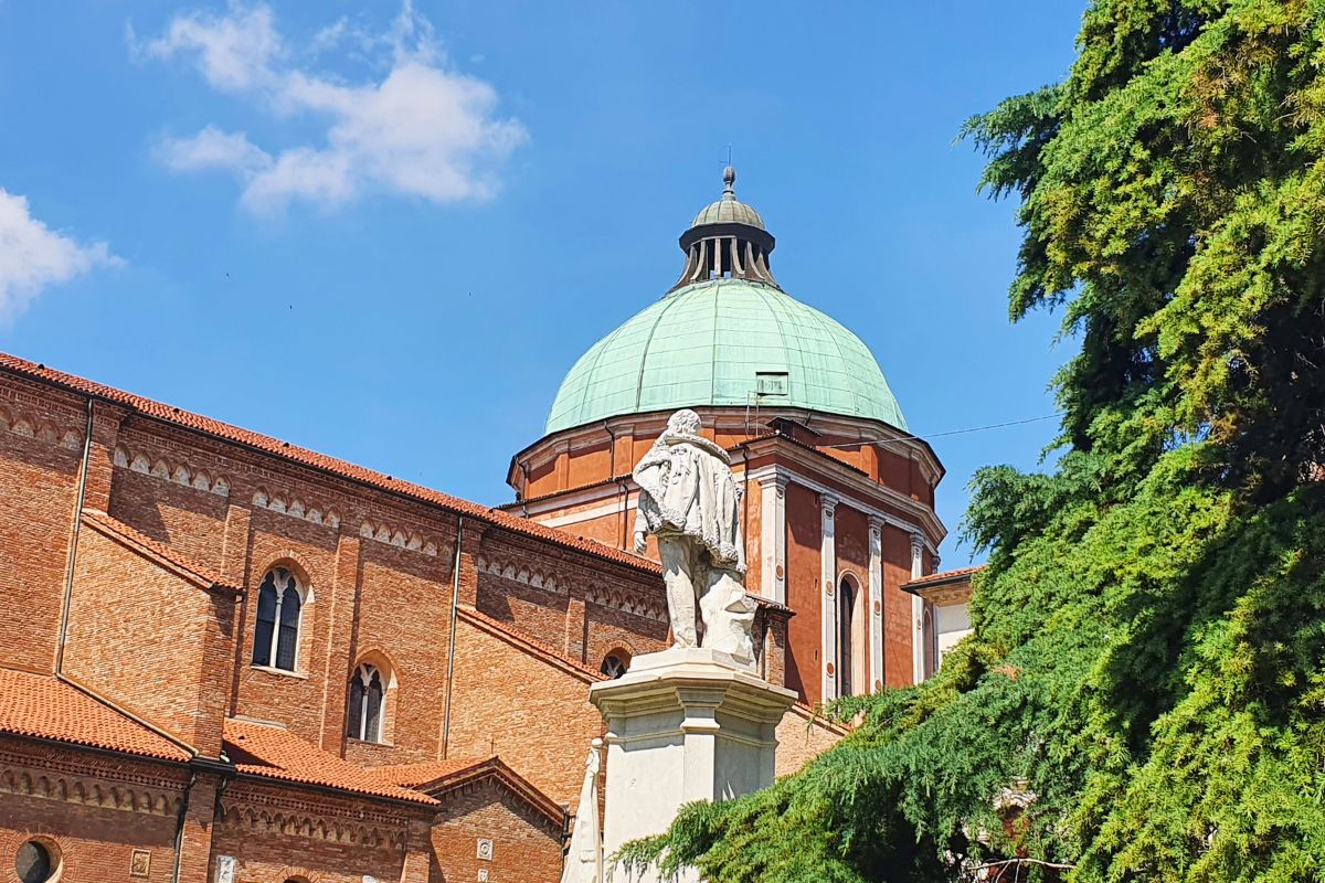 Blick über eine Statue (Rückenansicht) auf die massive Ziegelfassade und die markante grüne Kuppel der Kathedrale von Vicenza vor blauem Himmel.