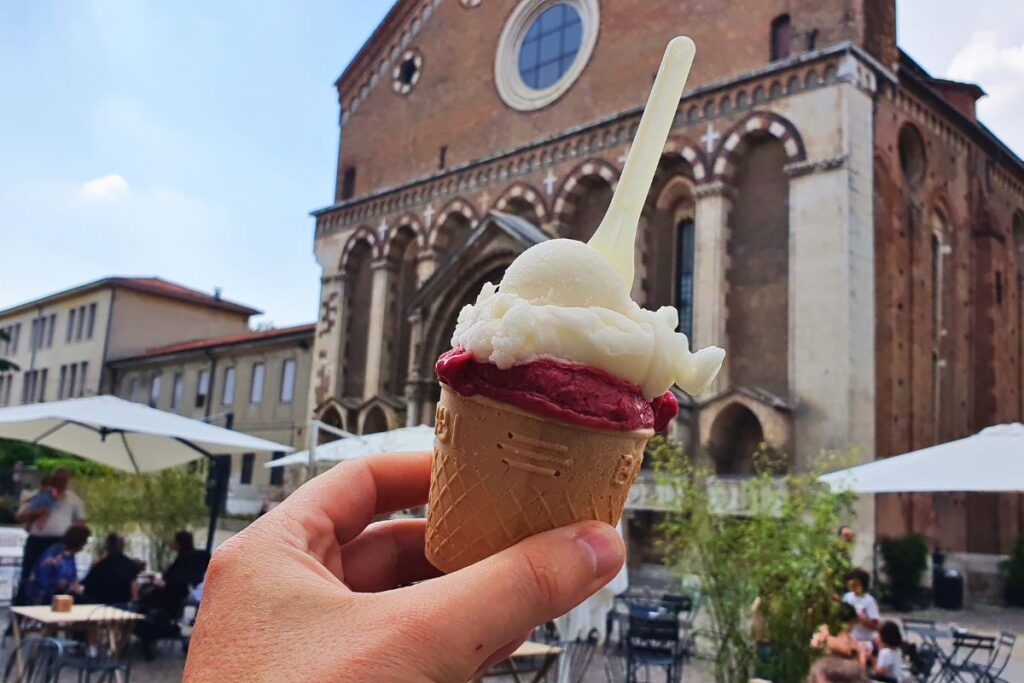 Eine Hand hält eine Waffel mit Frucht- und Milcheis im Vordergrund, unscharf im Hintergrund die Backsteinfassade der Kirche Tempio di San Lorenzo.