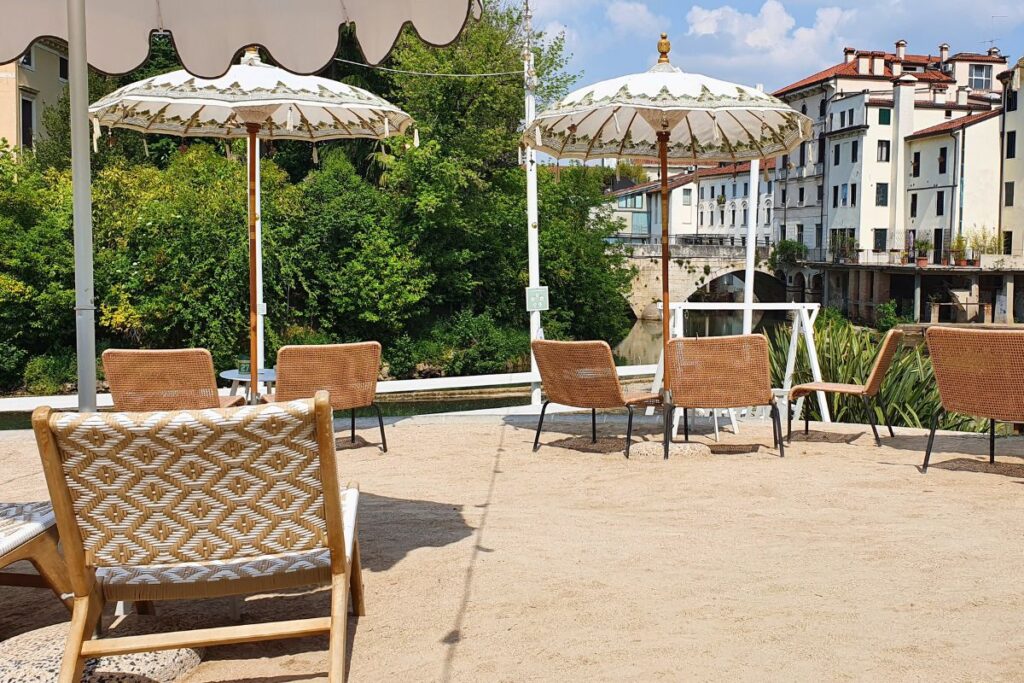 Entspannte Strandatmosphäre mitten in der Stadt: Bambus-Liegestühle und Sonnenschirme auf sandigem Boden in der Bar "Ultima Spiaggia" am Flussufer.