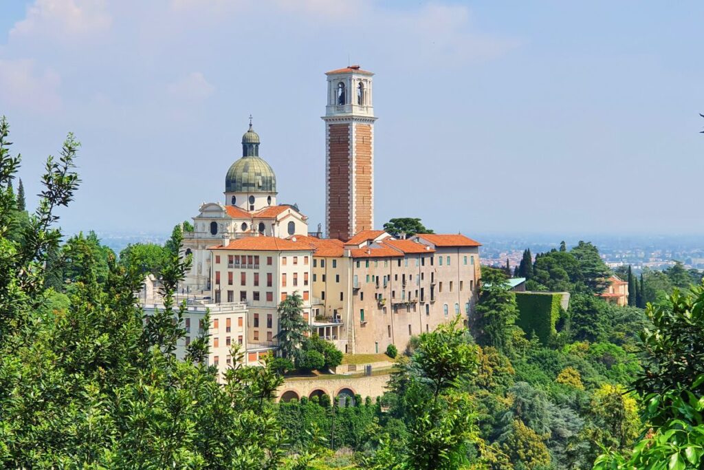 Die Basilika Monte Berico thront majestätisch auf einem grünen Hügel über Vicenza, flankiert von ihrem hohen Glockenturm vor blauem Himmel.