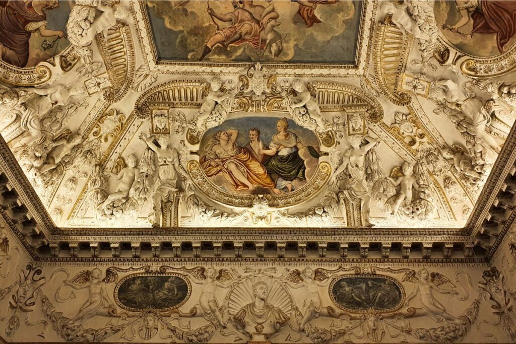 Eine überreich verzierte Zimmerdecke im Palazzo Barbaran mit goldenem Stuck, weißen Figurenreliefs und zentralen mythologischen Gemälden.