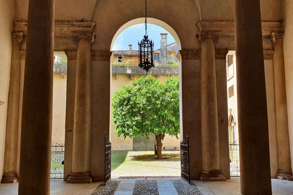 Blick durch massive Säulen in den sonnigen Innenhof des Palazzo Barbaran, in dessen Mitte ein grüner Baum steht.