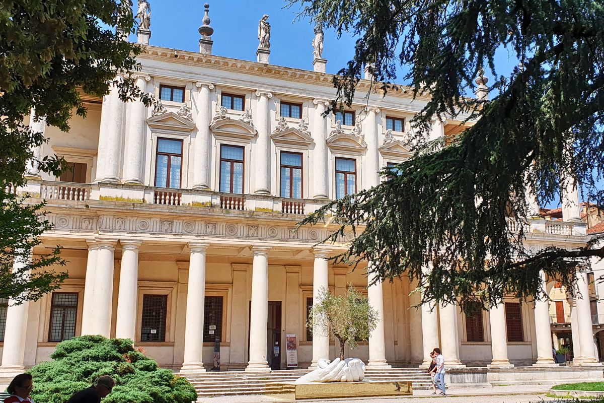 Die beeindruckende weiße Fassade des Palazzo Chiericati in Vicenza mit zwei übereinanderliegenden Säulenreihen (Loggien) und Statuen auf dem Dach, davor eine Grünfläche mit einer modernen Skulptur.