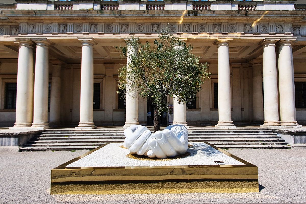 Die weiße Skulptur zweier riesiger Hände, die einen Olivenbaum halten, vor der imposanten Säulenfassade des Palazzo Chiericati unter blauem Himmel.