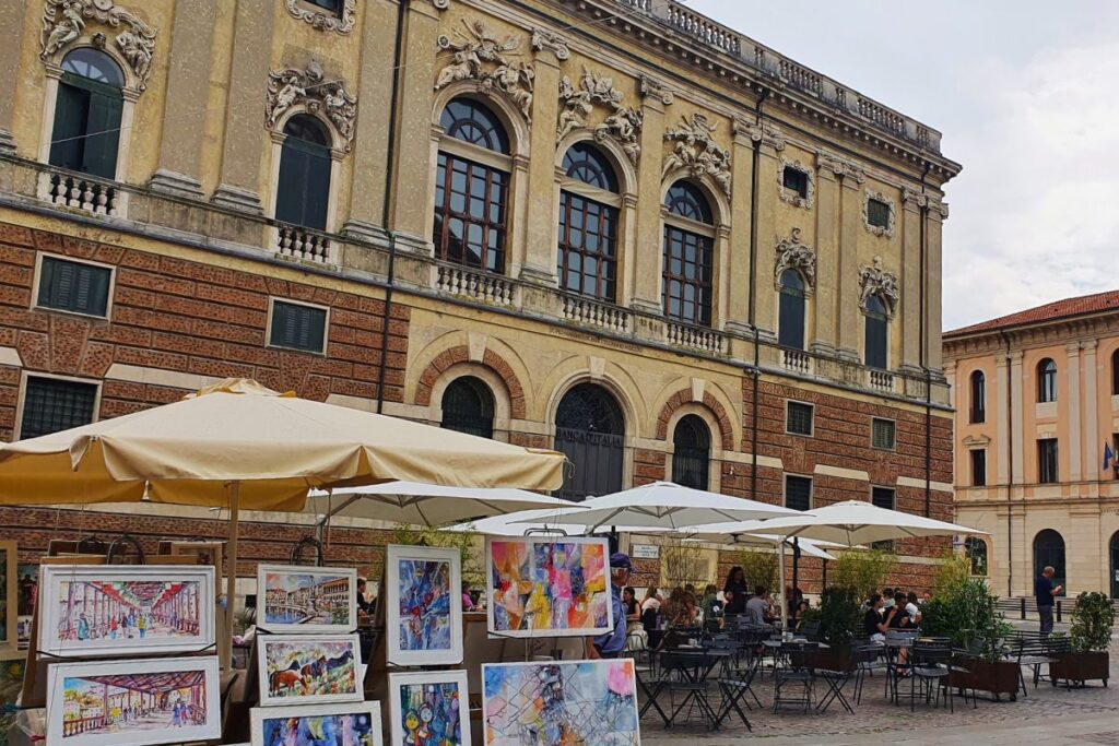 Die breite, ockerfarbene Fassade des Palazzo Repeta (heute Bank von Italien) mit großen Bogenfenstern, davor stehen Tische eines Cafés und Gemälde von Straßenkünstlern.