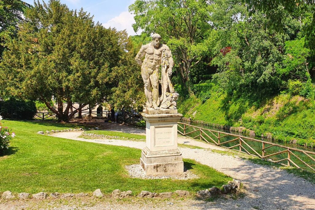 Eine verwitterte, klassische Steinstatue (Herkules) auf einem Sockel im Giardino Salvi, im Hintergrund viel Grün und ein kleiner Wasserlauf.