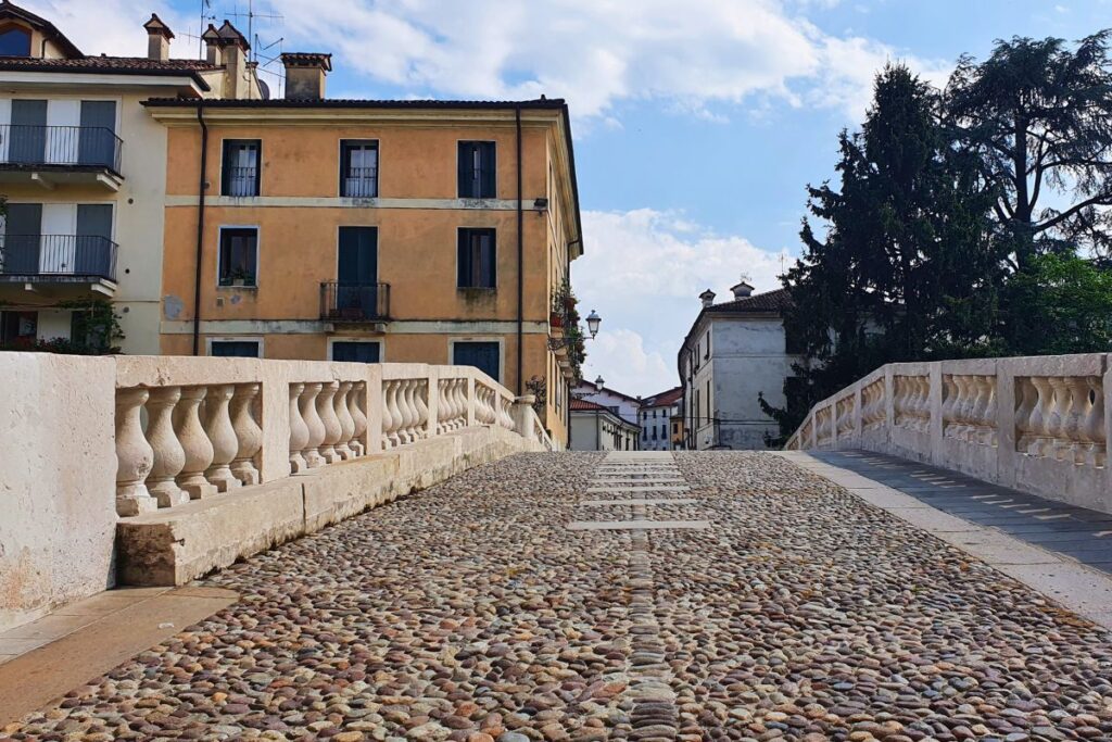Froschperspektive auf das historische Kopfsteinpflaster der Ponte San Michele in Vicenza. Die Brücke wird von hellen Steinbalustraden gesäumt und führt den Blick zu den alten Häusern am anderen Ufer.