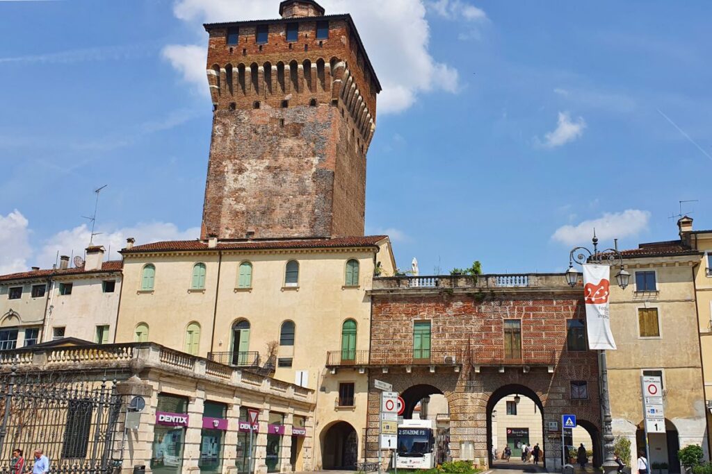 Der massive mittelalterliche Turm "Torrione" ragt hinter dem steinernen Bogen des Stadttors Porta Castello auf, daneben historische Gebäude unter blauem Himmel.