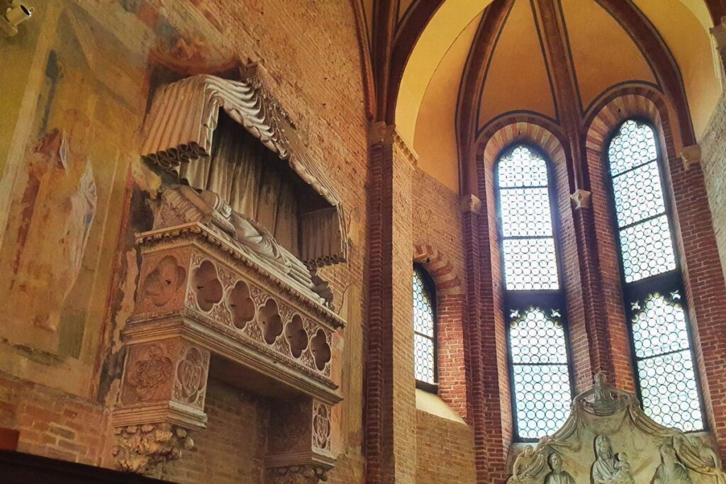 Ein historisches, an der Wand angebrachtes Grabmal mit steinernem Baldachin in der Kirche San Lorenzo, daneben hohe Fenster und Reste alter Wandmalereien.