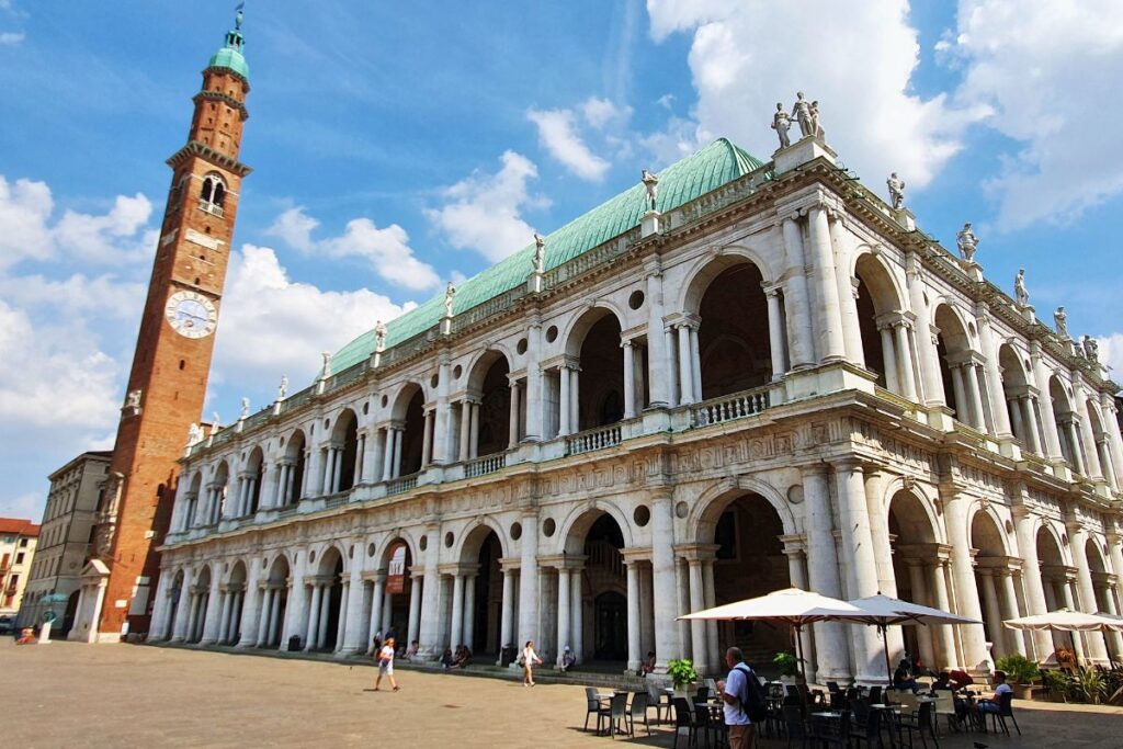 Die prachtvolle weiße Fassade der Basilica Palladiana mit ihren Säulengängen und dem grünen Kupferdach am Piazza dei Signori, daneben der hohe rote Torre Bissara unter blauem Himmel.