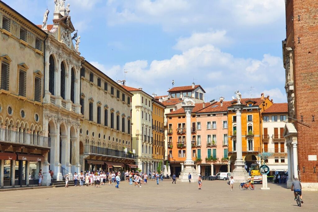 Weitwinkel-Ansicht der Piazza dei Signori in Vicenza mit der Basilica Palladiana, den venezianischen Säulen und Passanten unter blauem Himmel.