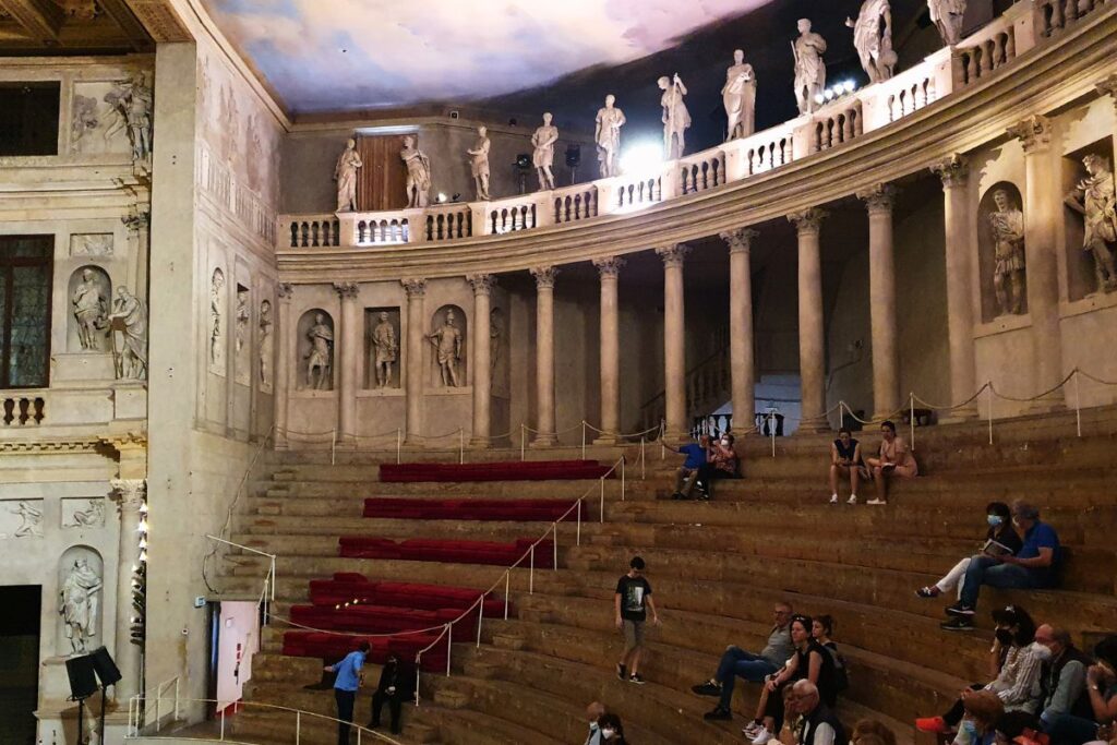 Der halbrunde Zuschauerraum (Cavea) des Teatro Olimpico mit steinernen Stufen, Säulengang mit Statuen oben und einer bemalten Decke, die einen wolkenverhangenen Himmel imitiert.