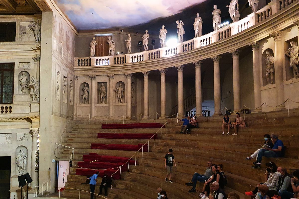 Der halbrunde Zuschauerraum (Cavea) des Teatro Olimpico mit steinernen Stufen, Säulengang mit Statuen oben und einer bemalten Decke, die einen wolkenverhangenen Himmel imitiert.