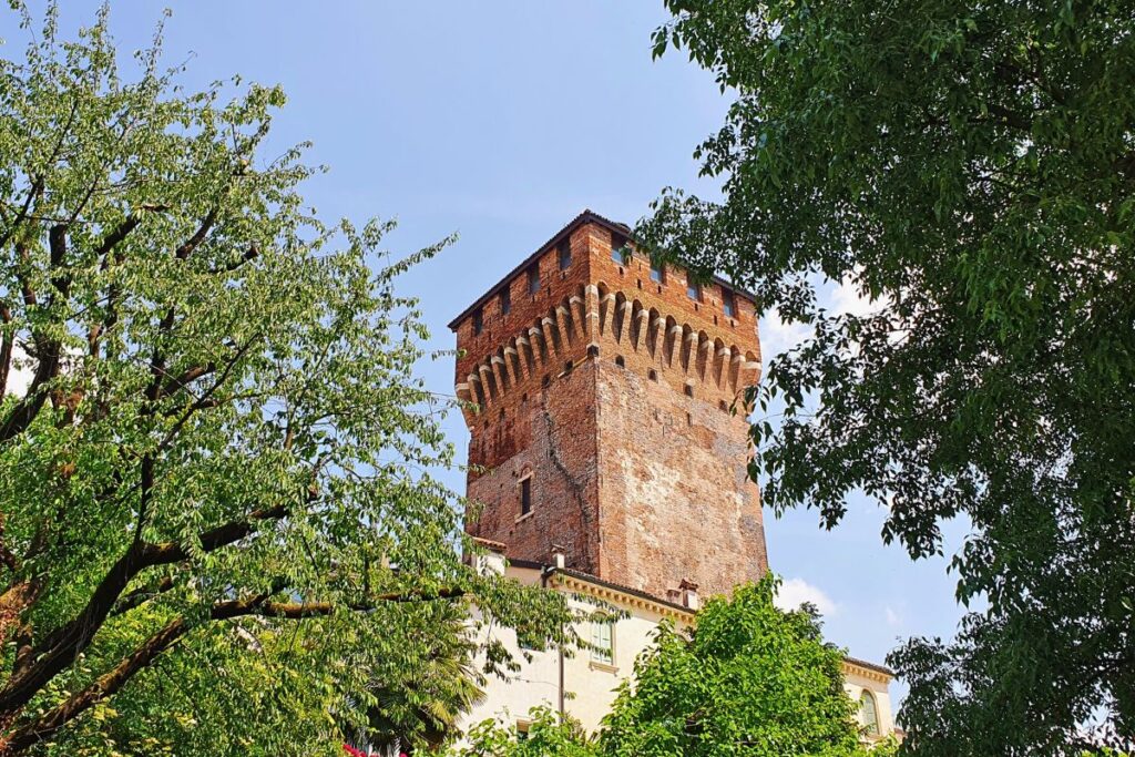Der wuchtige, mittelalterliche Wehrturm "Torrione" aus altem Backstein mit Zinnenkranz, umgeben von grünen Bäumen.