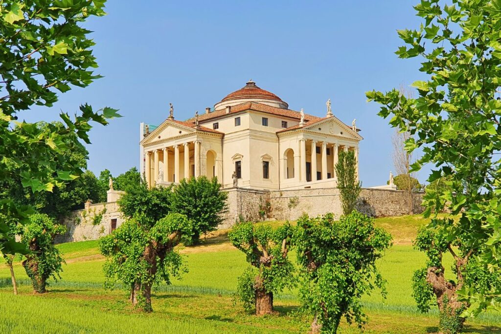 Die weltberühmte Villa La Rotonda (Villa Almerico Capra) auf einem grünen Hügel bei Vicenza. Zu sehen ist eine der vier identischen Tempelfronten mit sechs Säulen und Statuen auf dem Dach und der Treppe, umrahmt von Bäumen.
