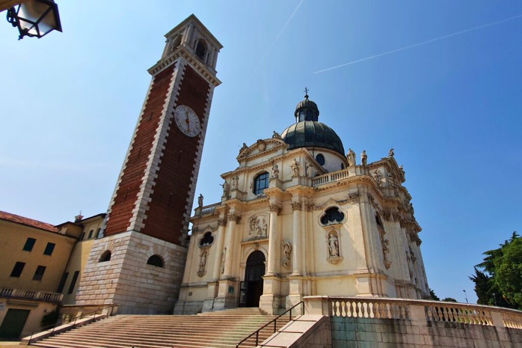 Froschperspektive der barocken Fassade der Wallfahrtskirche Monte Berico mit ihren Statuen und der Kuppel, links daneben ragt der rote Glockenturm auf.
