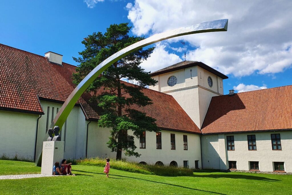 Das weiße Gebäude des Wikingerschiffsmuseums (Vikingskipshuset) mit rotem Ziegeldach und einer modernen Metallskulptur im Garten