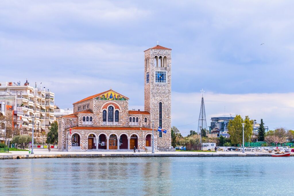 Die hellbraune Steinkirche Agios Konstantinos und Eleni mit ihrem hohen Glockenturm direkt an der Uferpromenade von Volos unter einem weiten Himmel.