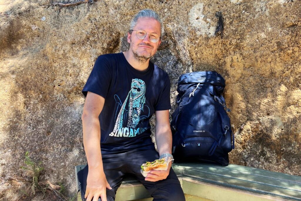 Nicolo Martin sitzt entspannt mit einem blauen Tagesrucksack an einer Felswand und isst ein Sandwich.