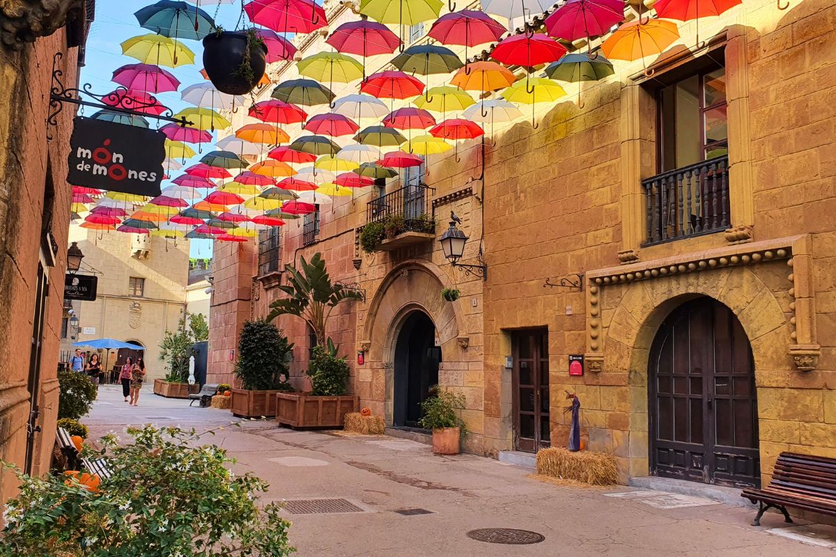 Eine charmante Gasse im Poble Espanyol in Barcelona mit hunderten farbenfrohen Regenschirmen, die als Sonnenschutz über der Straße hängen.
