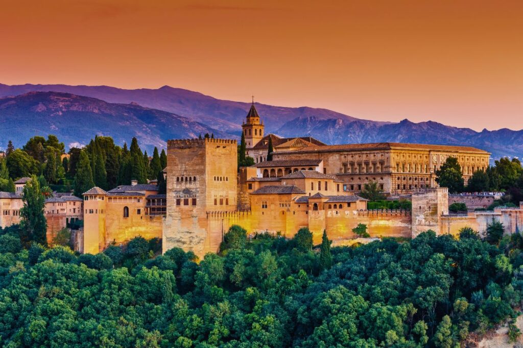 Die historische Festungsanlage Alhambra in Granada vor schneebedeckten Bergen im warmen, orangefarbenen Licht des Sonnenuntergangs.